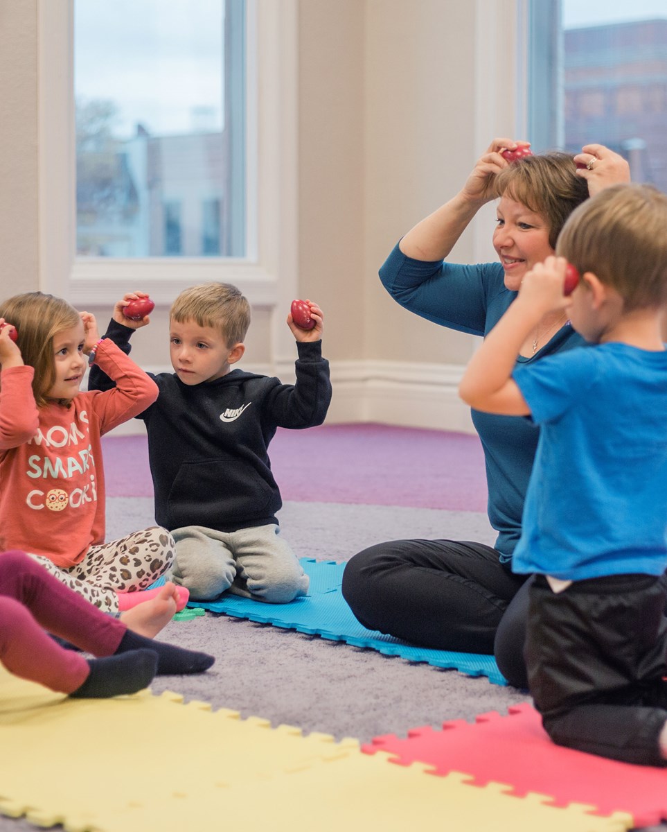 A teacher sits with students on the floor and demonstrates how to shake egg shakers from Kindermusik’s childcare resources kit.